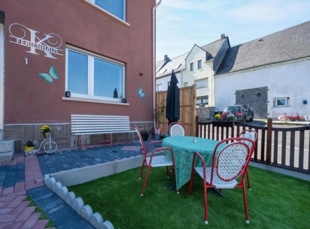 Terrasse/Gartenbereich vor der Hausfassade mit 'Ferienhaus'-Schriftzug, Sitzgruppe mit rot-weißen Stühlen auf Kunstrasen, im Hintergrund Nachbargebäude und parkende Autos an der Straße sichtbar.