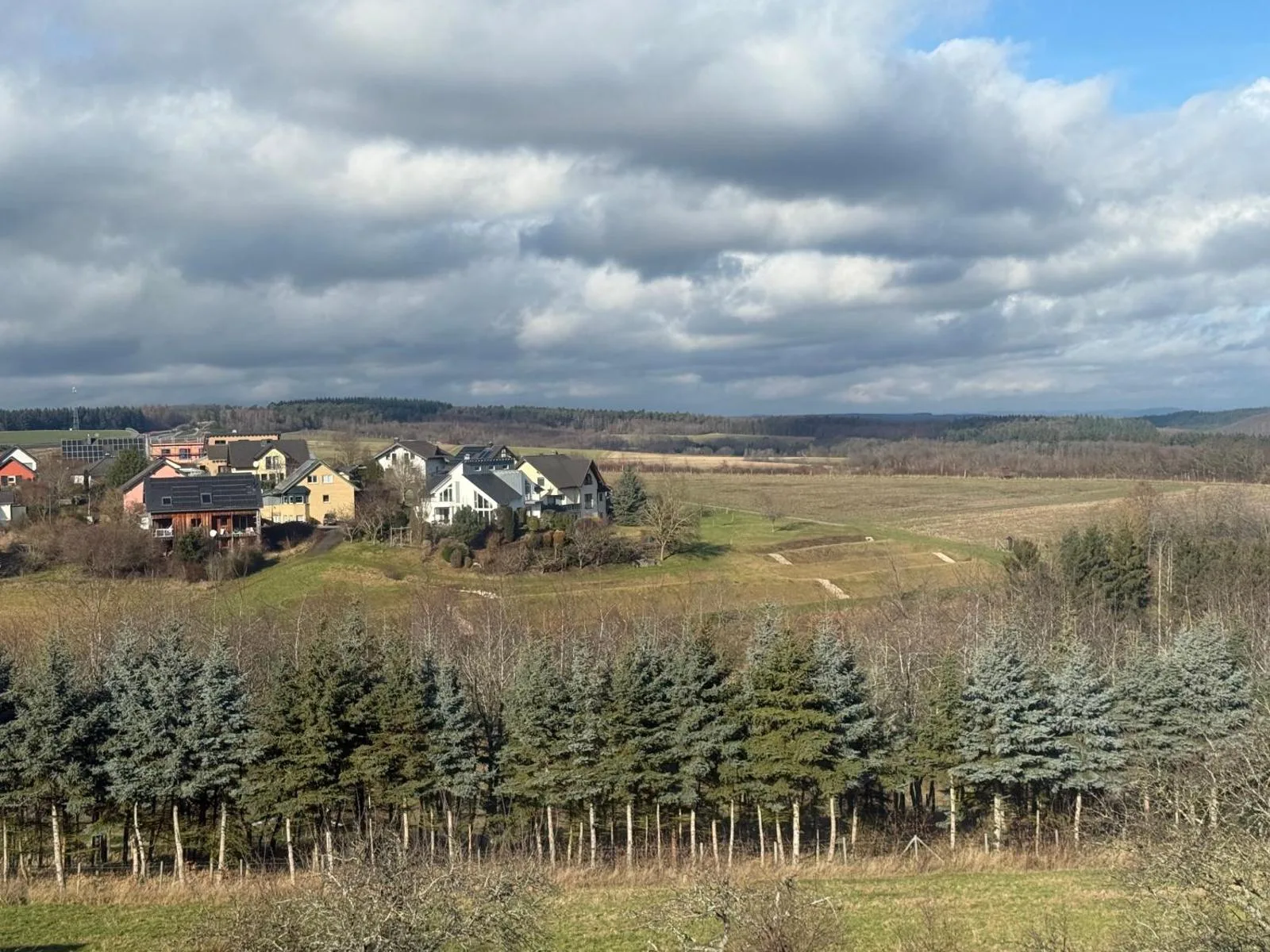 Panoramablick über das Dorf Longkamp mit mehreren Häusern, Nadelbaumreihe im Vordergrund und dramatischem Wolkenhimmel im Hintergrund.