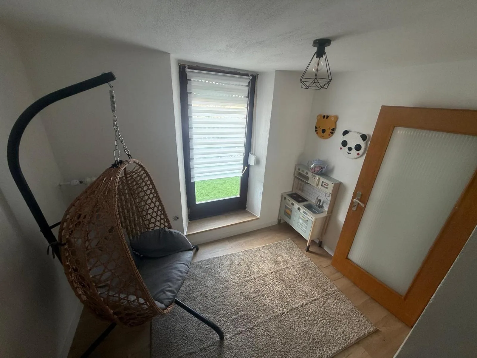 Kinderzimmer mit hängendem Rattan-Sessel, Fenster mit Gartenblick, Holz-Spielküche und Tiger/Panda-Wandfilzbildern.