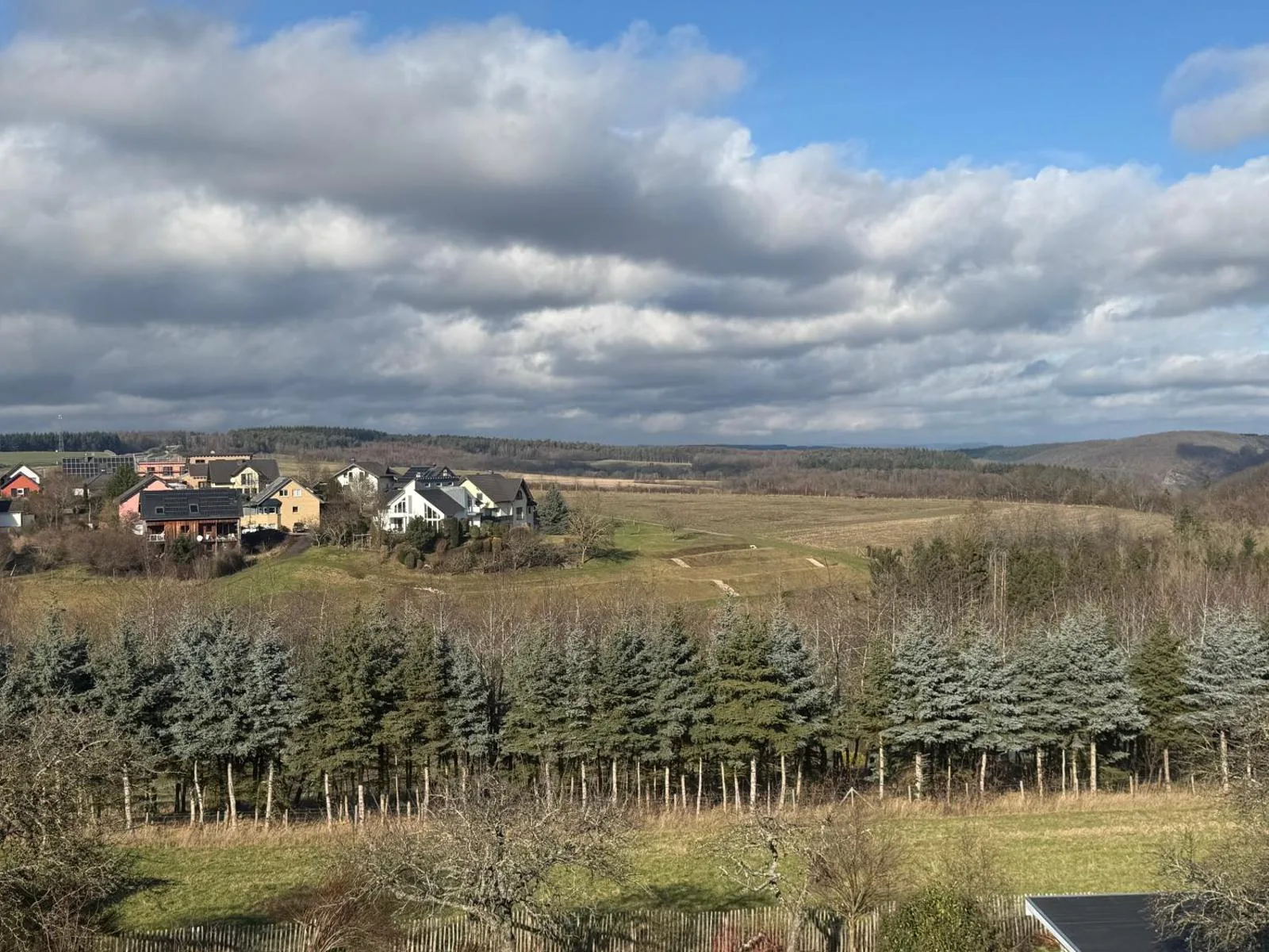 Blick über Dorf Longkamp mit mehreren Häusern und Nadelbaumreihe bei strahlend blauem Himmel (Tageszeit-Duplikat von Bild 01).