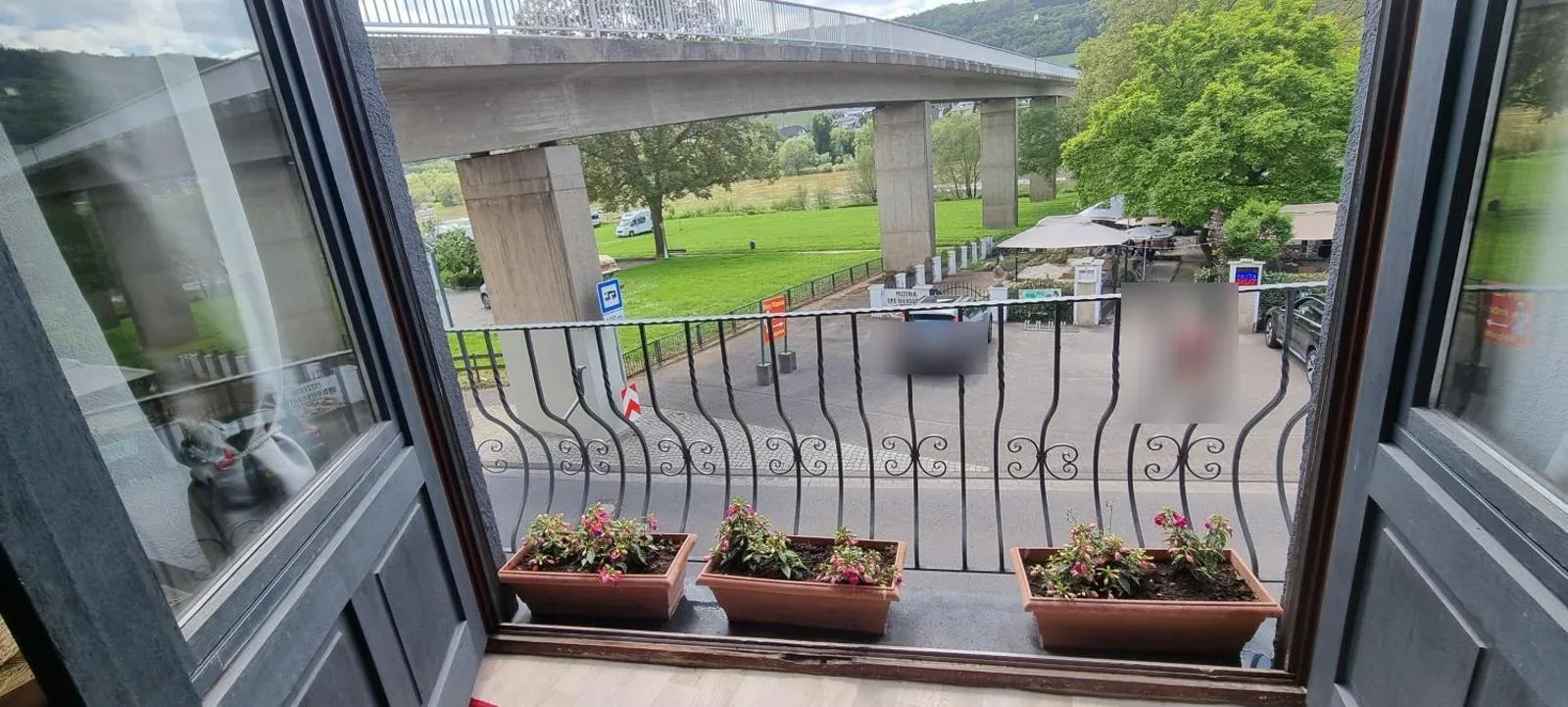 Blick durch geöffnete Balkontür mit Blumenkästen auf Brücke, Parkplatz, Restaurantterrasse und Grünanlage