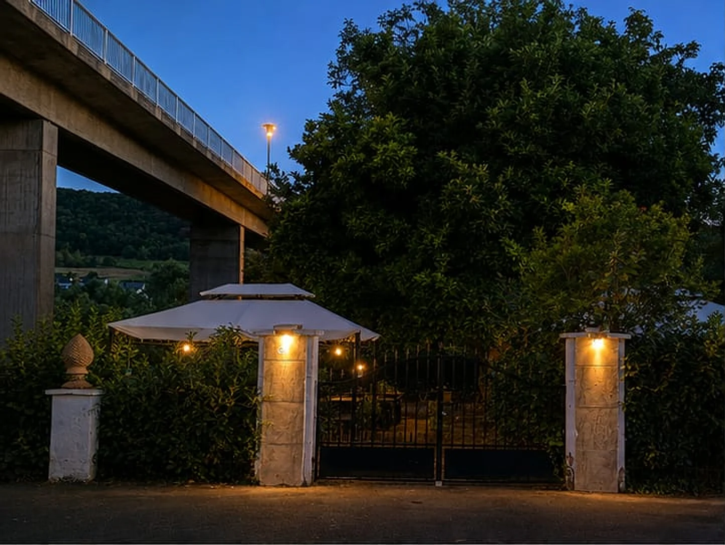 Beleuchtete Einfahrt mit Torpfeilern und Sonnenschirmen in der Abenddämmerung, dahinter die Moselbrücke