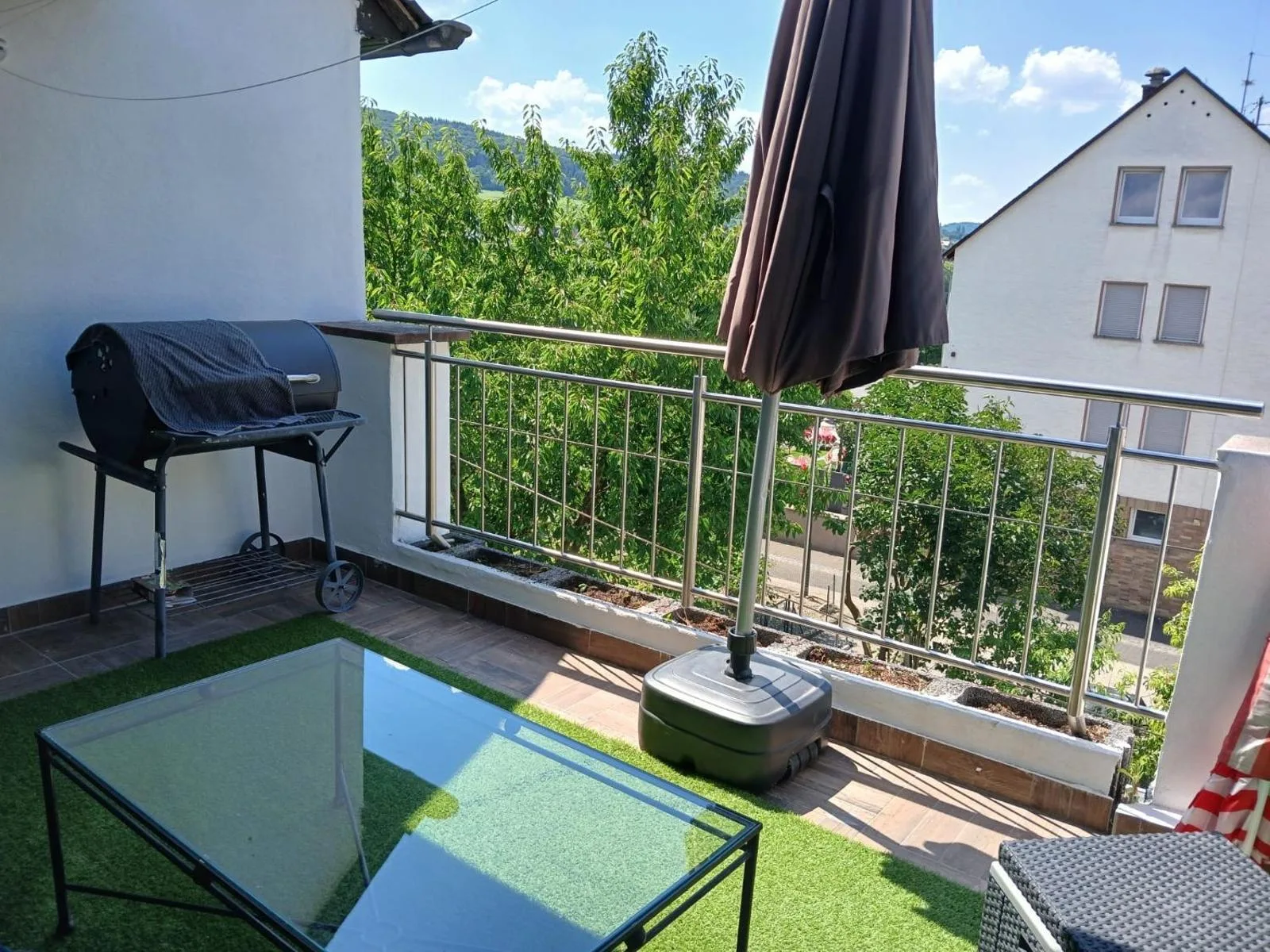 Balkon mit Holzkohlegrill, Glastisch auf Kunstrasen und aufgespanntem Sonnenschirm, Blick ueber gruene Baeume und Huegel bei blauem Himmel.