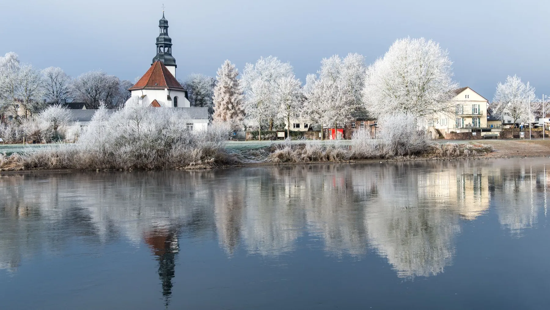Raureif an einem Flussufer mit Kirchturm im Winter